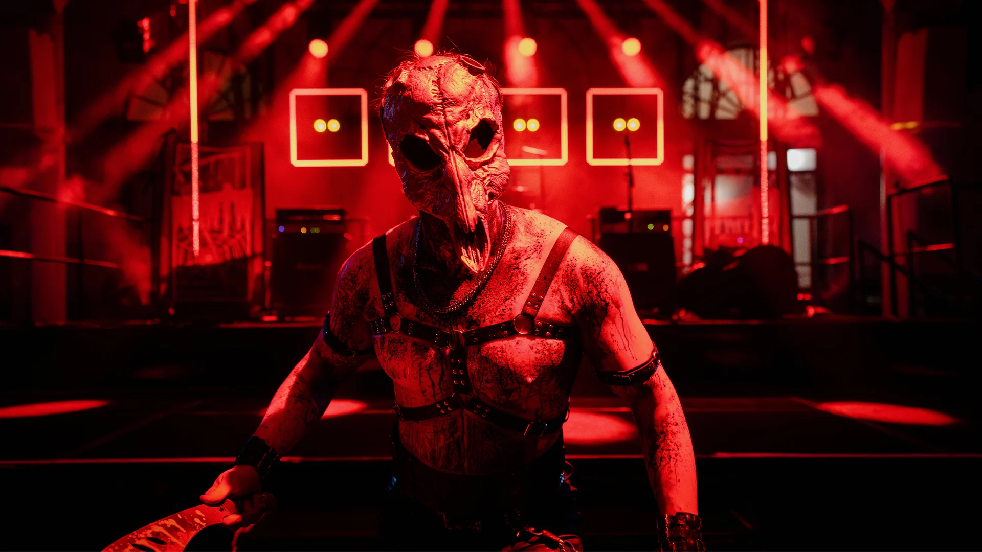 A person in a menacing bird skull mask stands shirtless in red stage lighting, holding a weapon, with a concert stage and bright lights behind them.