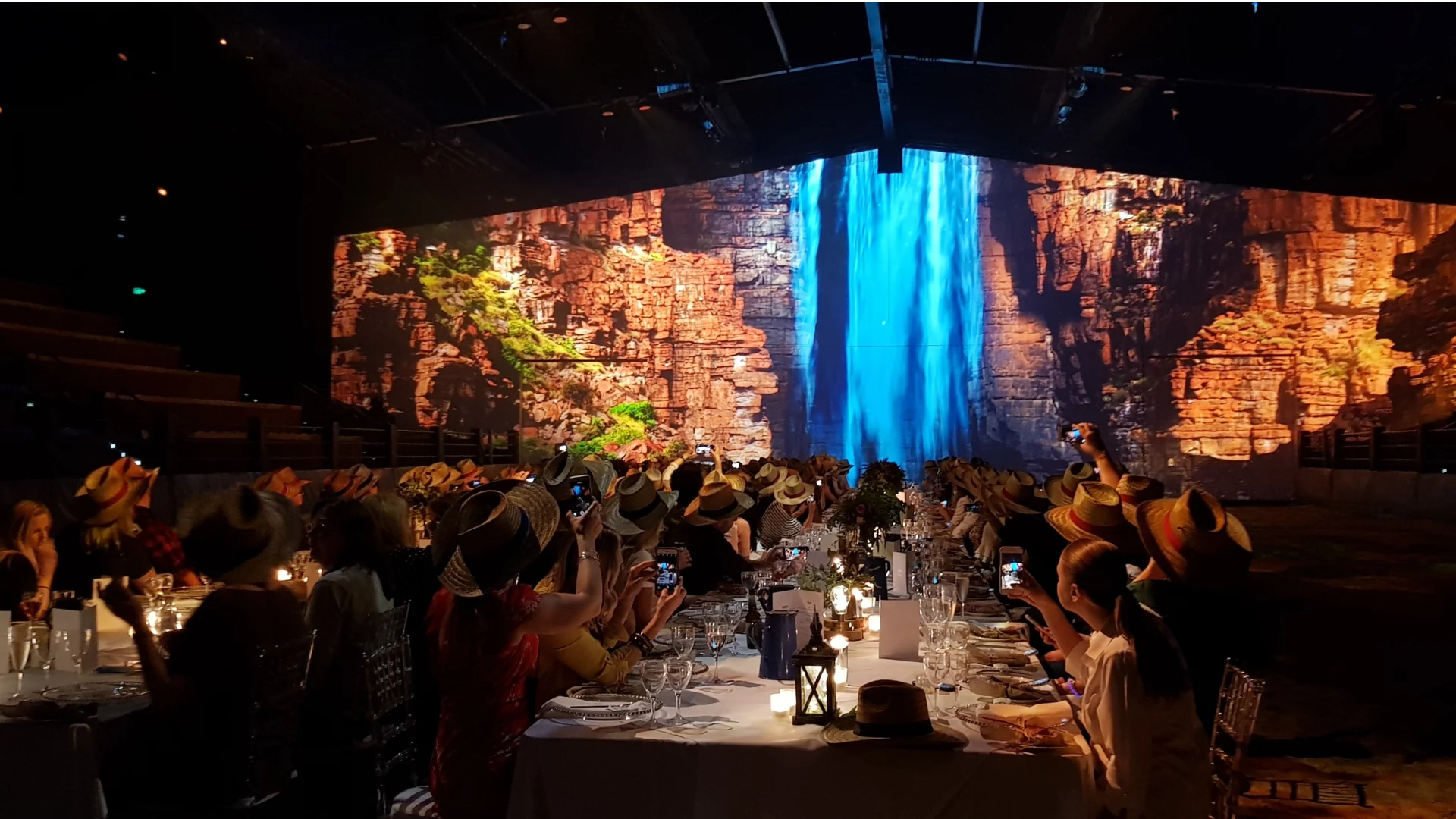 A large group of people wearing hats sit at long, elegantly set tables, facing a stage with a vivid backdrop of a waterfall cascading down rocky cliffs, under dim, dramatic lighting.