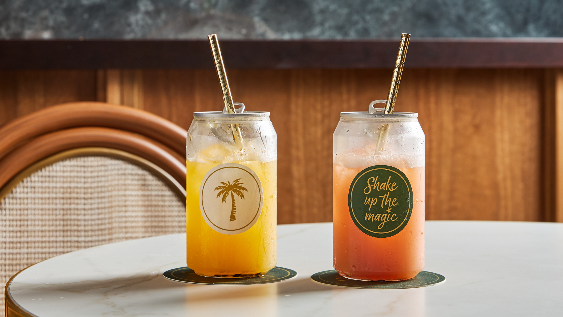 Two iced drinks in clear cans with straws sit on a round white table. The left drink is yellow-orange with a palm tree graphic; the right is pink-orange with a “Shake up the magic” label. Both rest on green coasters.