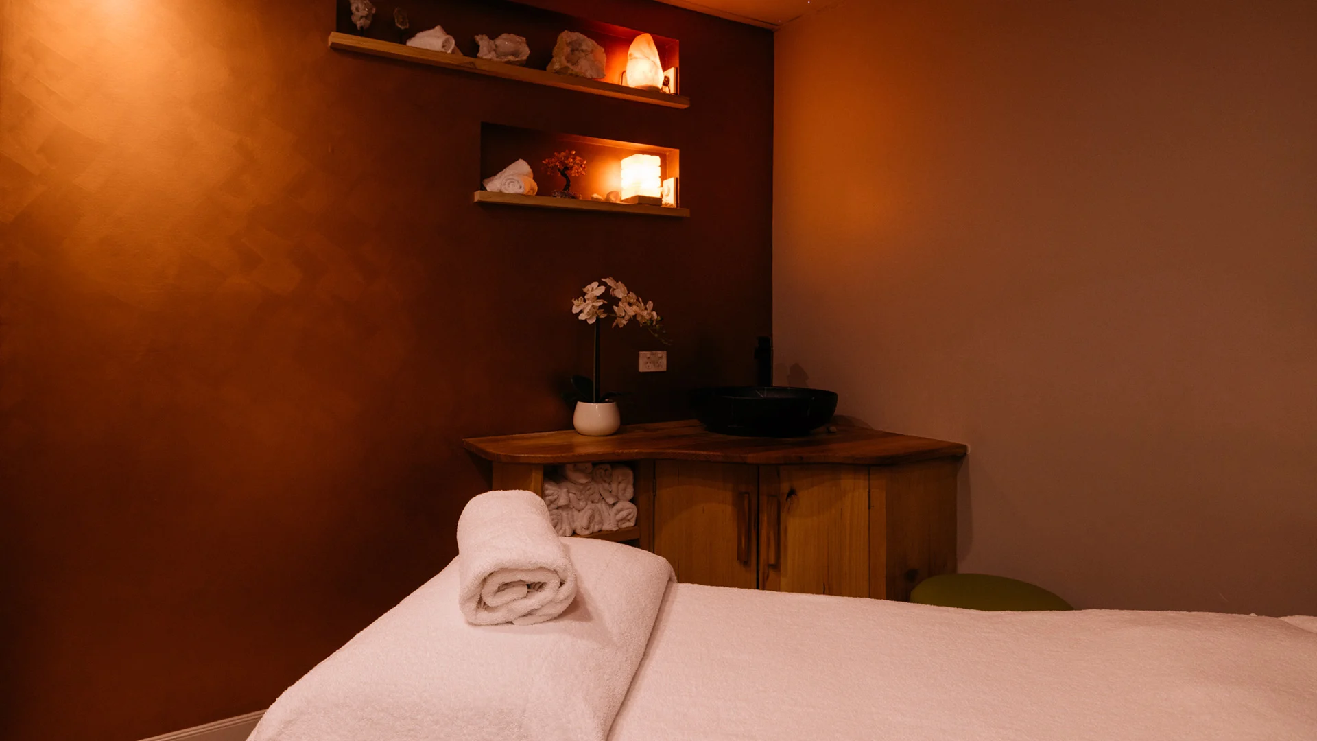 A serene spa room with a massage table, rolled towel, wooden cabinets, and softly lit shelves displaying candles and decorative items.
