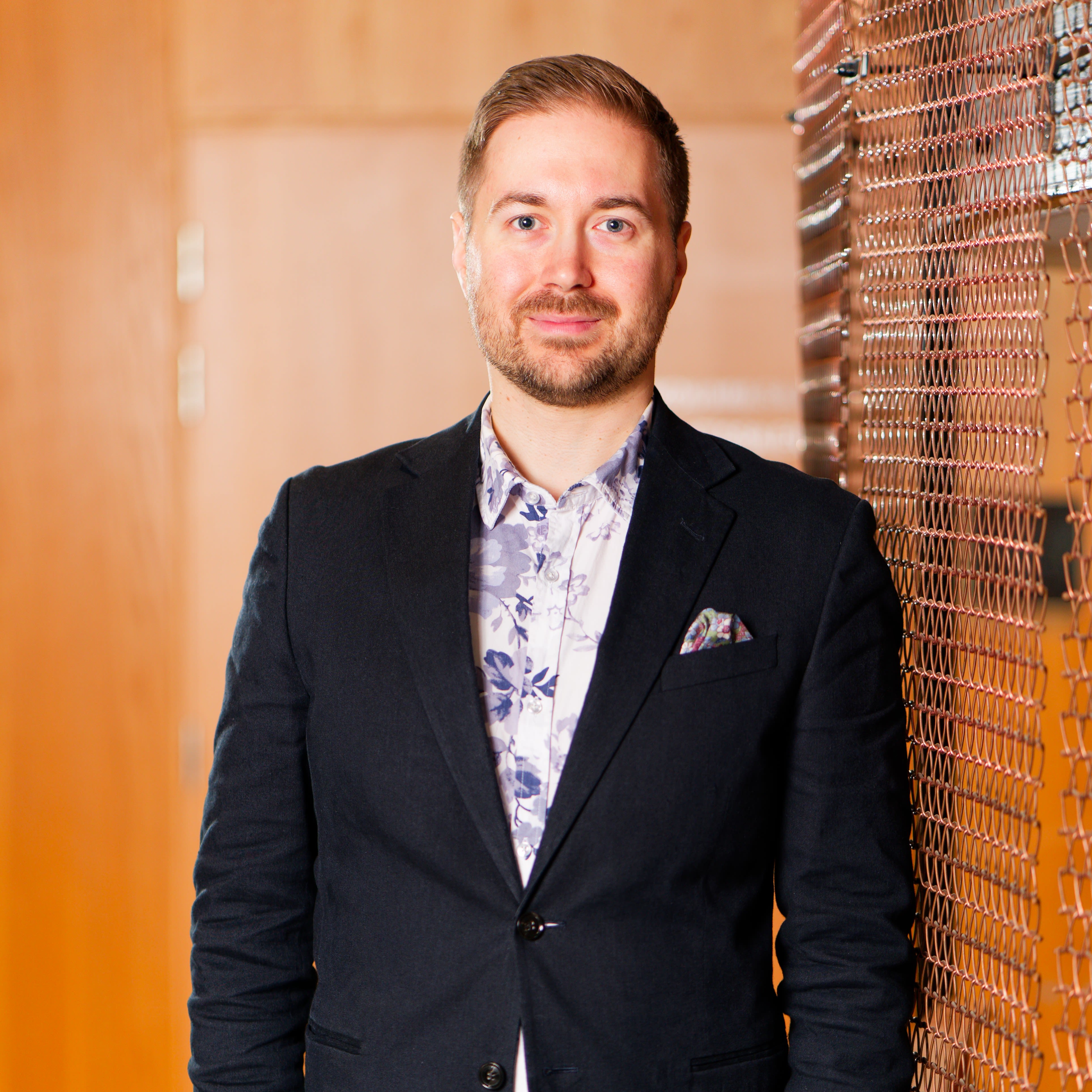 Lauri Lavanti seisomassa. Päällään hänellä on musta bleiseri ja sinivalkoinen kukkakuvioinen kauluspaita. Bleiserin taskussa on monivärinen taskuliina. Taustalla puunruskea käytävä.