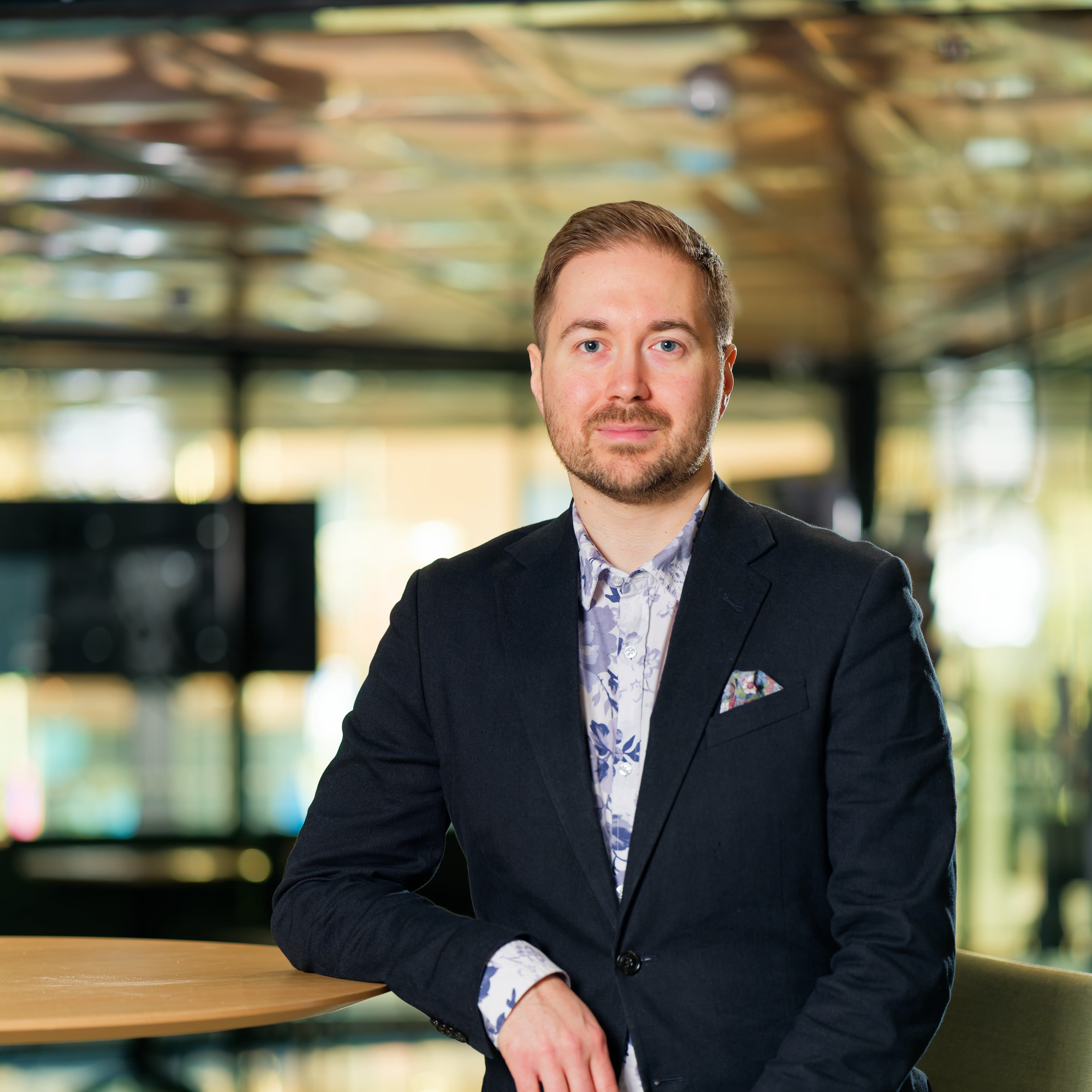 Lauri Lavanti nojaamassa korkeaan pöytään. Päällään hänellä on musta bleiseri ja sinivalkoinen kukkakuvioinen kauluspaita. Bleiserin taskussa on monivärinen taskuliina. Taustalla ikkunoita.