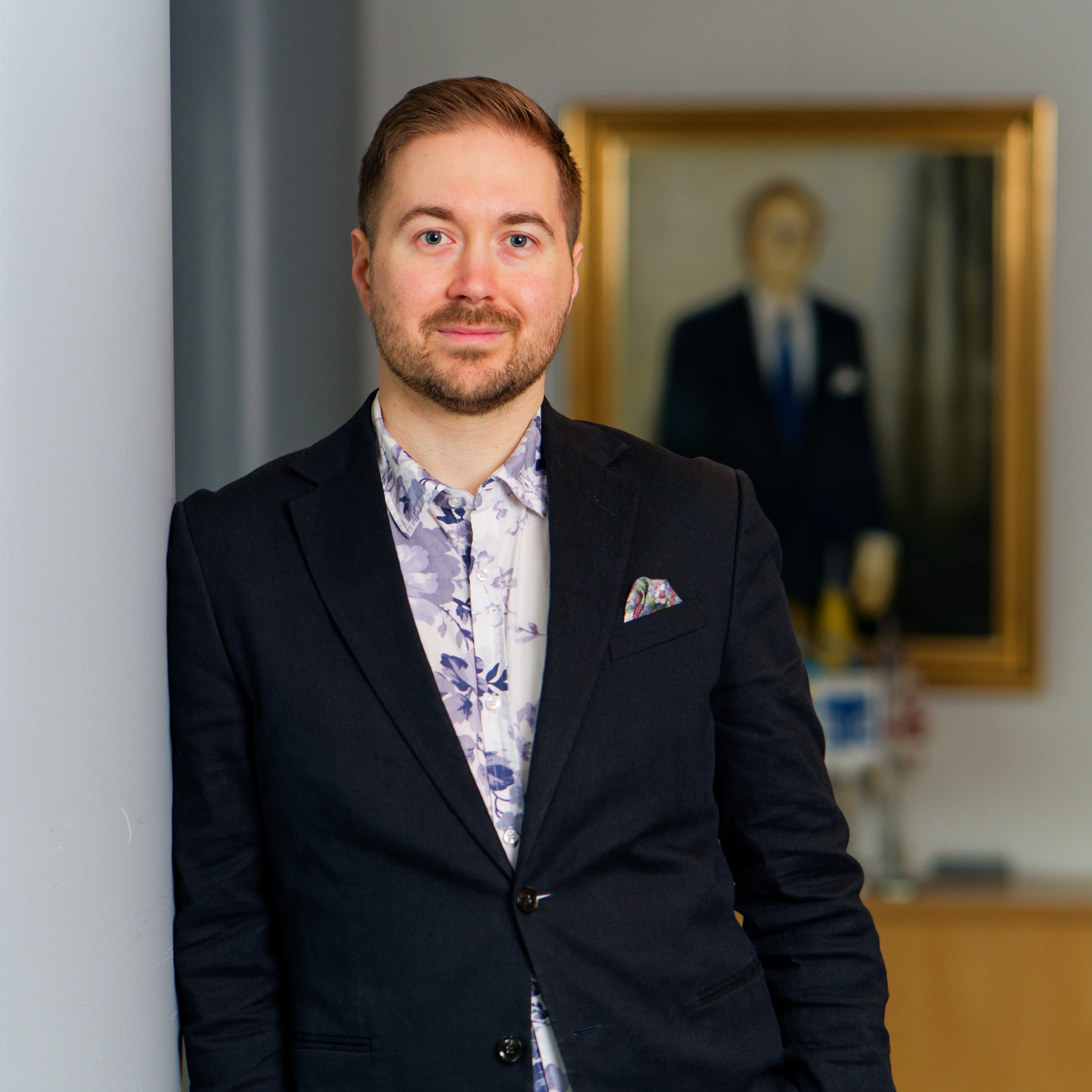 Lauri Lavanti nojaamassa pylvääseen. Päällään hänellä on musta bleiseri ja sinivalkoinen kukkakuvioinen kauluspaita. Bleiserin taskussa on monivärinen taskuliina. Taustalla taulu.