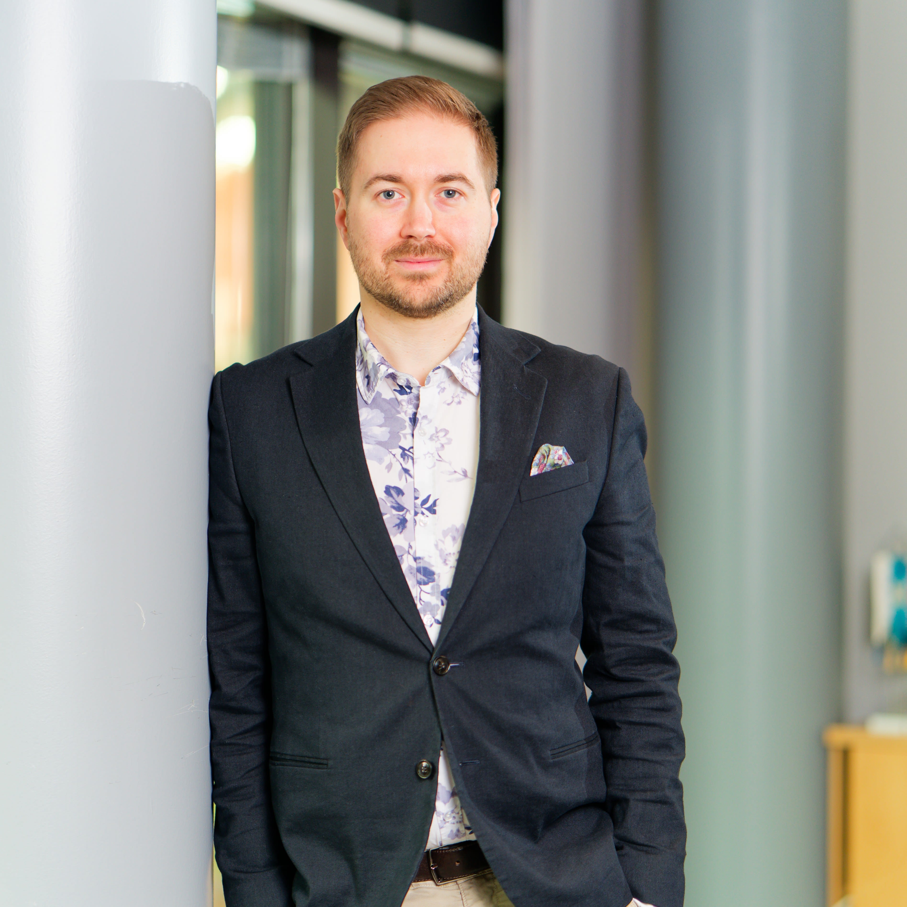 Lauri Lavanti seisomassa ja nojaamassa pylvääseen. Päällään hänellä on musta bleiseri ja sinivalkoinen kukkakuvioinen kauluspaita. Bleiserin taskussa on monivärinen taskuliina. Taustalla ikkunoita.