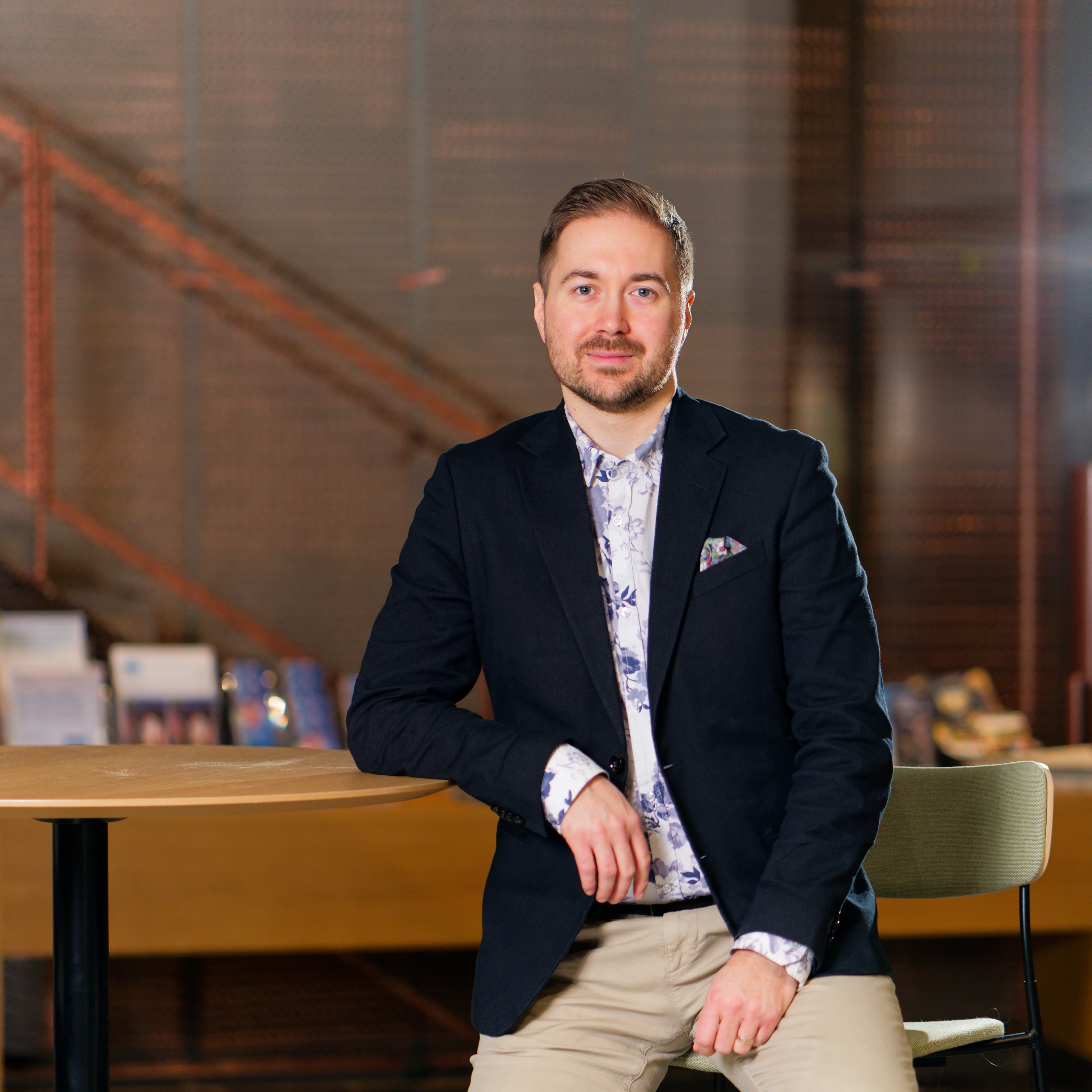 Lauri Lavanti istumassa jakkaralla ja nojaa korkeaan pöytään. Päällään hänellä on musta bleiseri ja sinivalkoinen kukkakuvioinen kauluspaita. Bleiserin taskussa on monivärinen taskuliina. Taustalla portaikko.