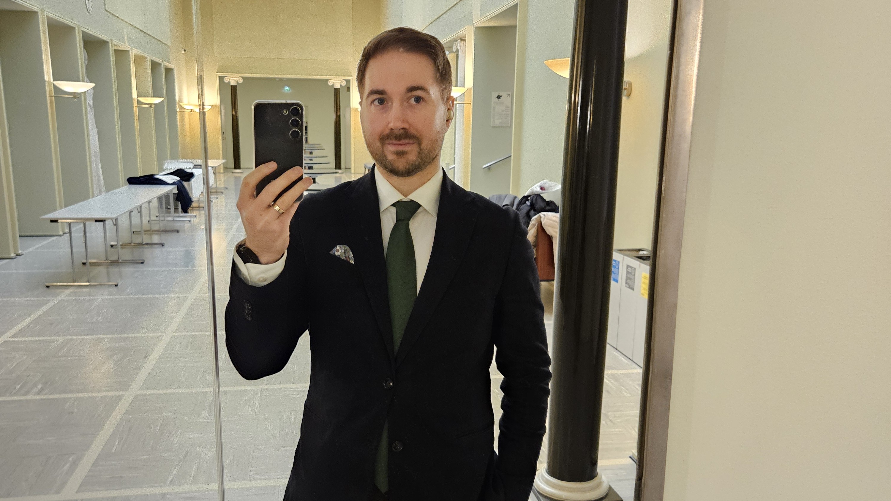 Lauri Lavanti seisomassa peilin edessä puhelin kädessä, ottamassa itsestään kuvaa. Päällä musta takki, valkoinen kauluspaita ja kaulassa vihreä kravatti. Taustalla Helsingin Yliopiston päärakennuksen vaalea käytävä.