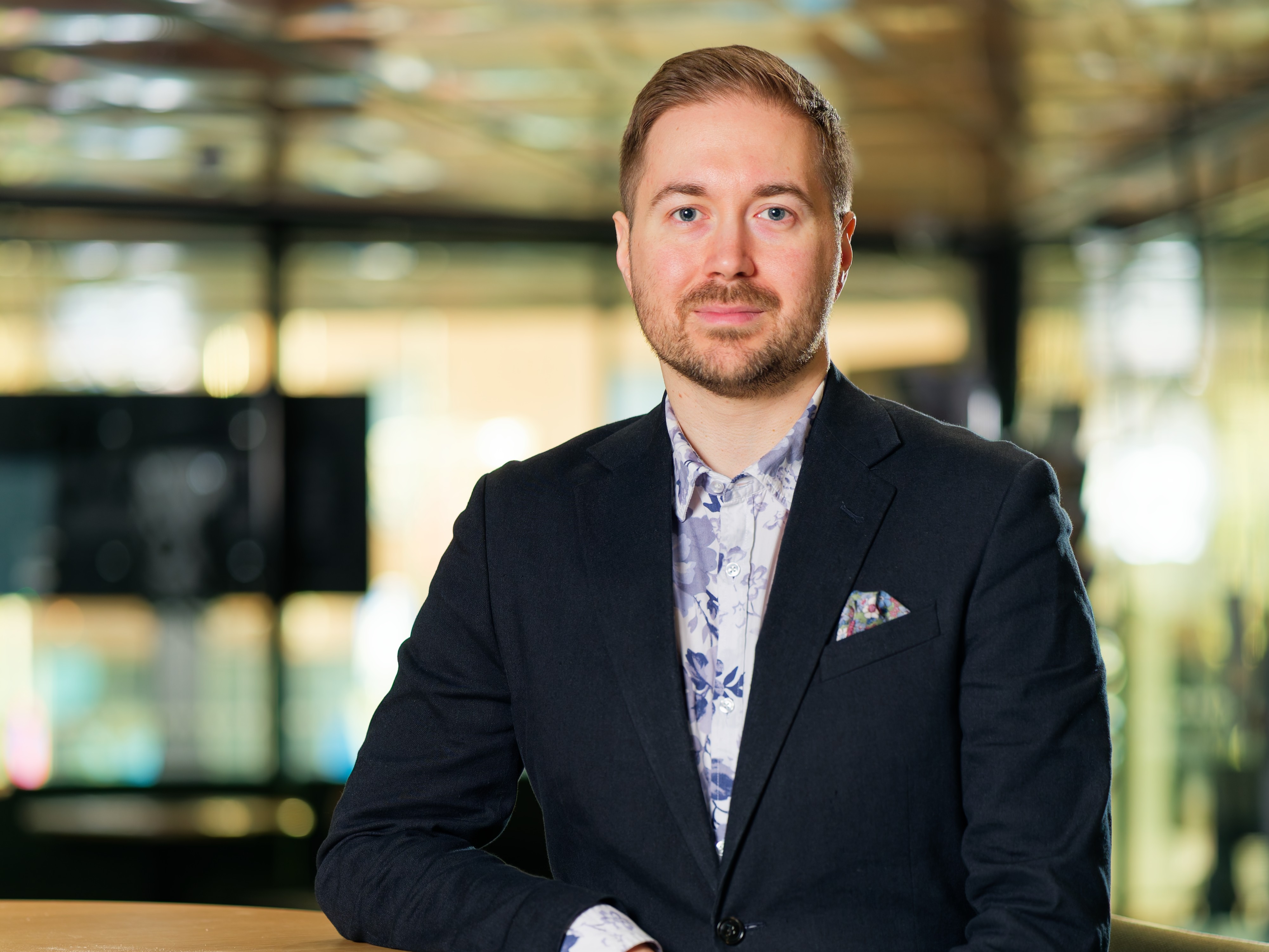 Lauri Lavanti nojaamassa korkeaan pöytään. Päällään hänellä on musta bleiseri ja sinivalkoinen kukkakuvioinen kauluspaita. Bleiserin taskussa on monivärinen taskuliina. Taustalla ikkunoita.