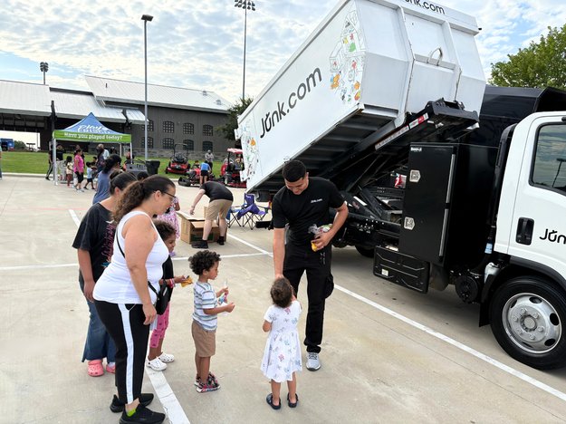 Junk.com truck arrives at Touch-A-Truck event to dazzle children. A junk crew member passes out plush trucks while the junk truck shows off its motorized dumping ability in the background.