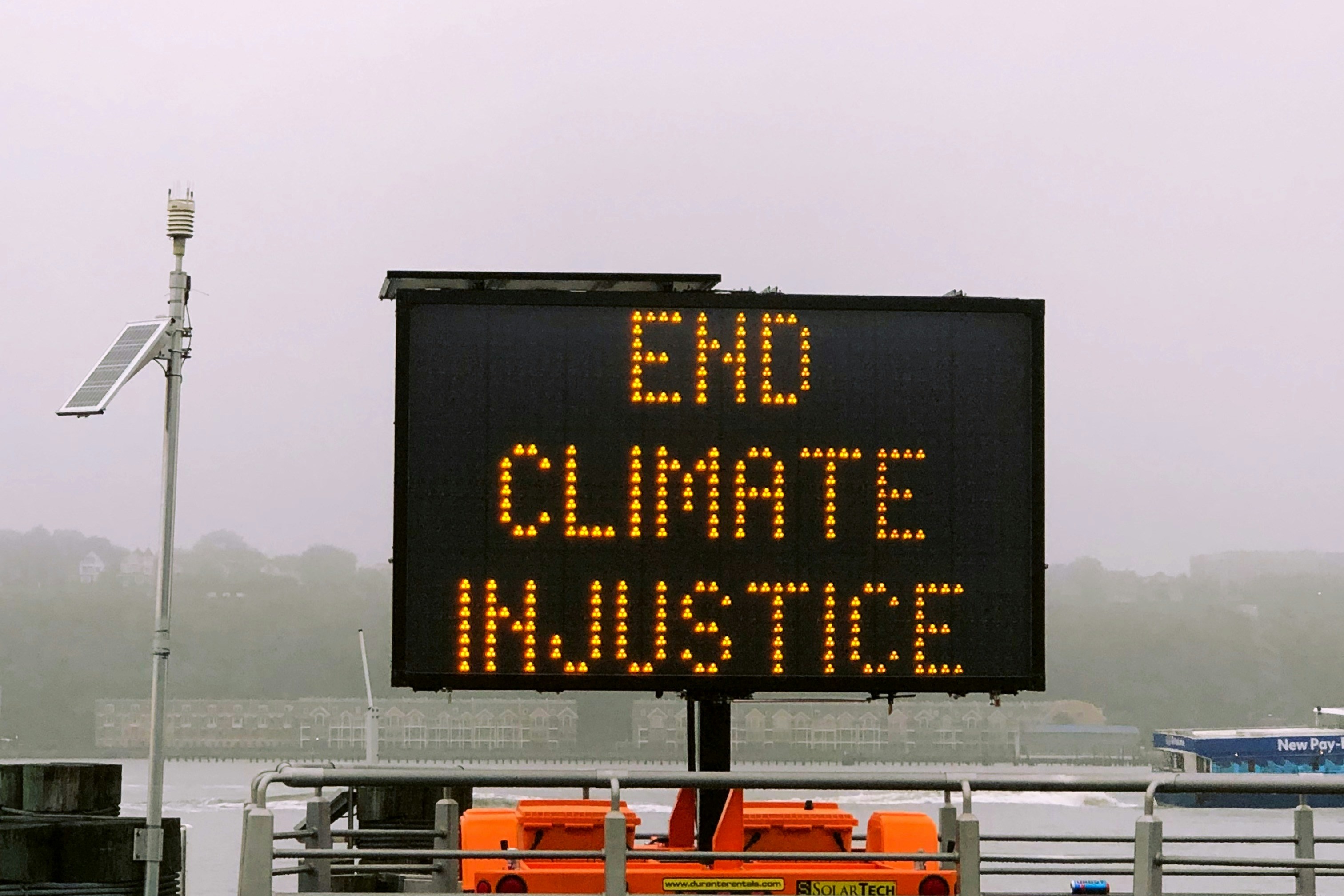 An electronic sign with bright yellow text reads END CLIMATE INJUSTICE against a foggy background. The sign is mounted near a railing with a solar panel and orange equipment nearby.