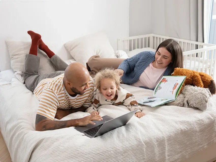 Kuva nauravasta lapsesta joka makaa vatsallaan sängyllä vanhempiensa kanssa ja selaa kannettavaa tietokonetta.