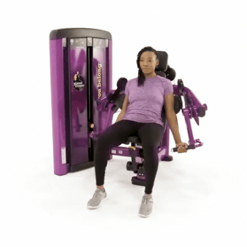 Woman sitting on Planet Fitness biceps curl machine and lifting arm to perform exercise