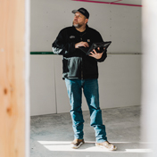 A man in a black jacket and cap holds a tablet, looking up and to the side inside a room with unfinished drywall.