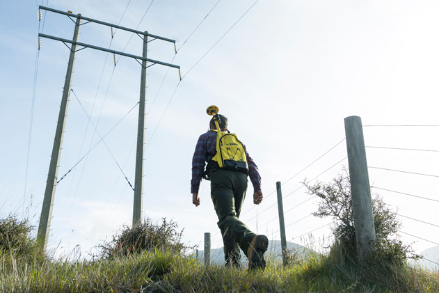 A person with a bright yellow. Trimble backpack and surveying equipment walks up a grassy hill towards large power lines and utility poles against a clear sky.