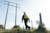A person with a bright yellow. Trimble backpack and surveying equipment walks up a grassy hill towards large power lines and utility poles against a clear sky.