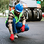 A construction worker in a blue hard hat and red gloves crouches to inspect fresh asphalt near a road roller on a paving site.