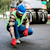A construction worker in a blue hard hat and red gloves crouches to inspect fresh asphalt near a road roller on a paving site.