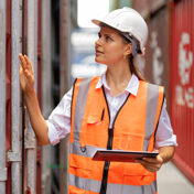 A female construction worker with protective equipment uses a tablet while inspecting red shipping containers.