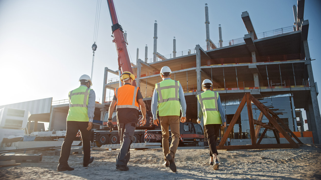 Four construction workers in hard hats and reflective vests walk towards a building under construction, with a large crane in the background.
