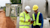 Two construction workers, wearing hard hats and safety vests, look at a digital map on a tablet showing a road and a blue line representing a utility.
