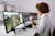 A woman with curly hair sits at a desk, looking at three computer monitors displaying 3D models and data visualizations.