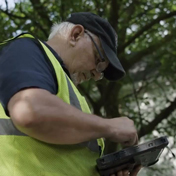 A man in a baseball cap and safety vest is focused on a handheld tablet or device.