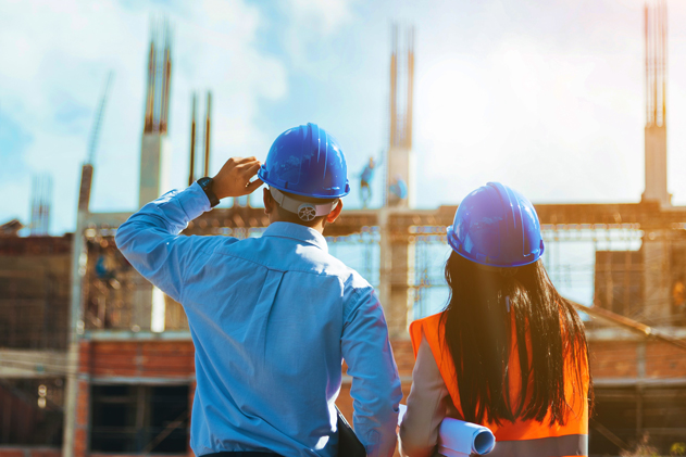 Two individuals in blue hard hats and safety vests stand with their backs to the viewer, observing a construction site with partially built structures under a bright sky.