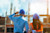 Two individuals in blue hard hats and safety vests stand with their backs to the viewer, observing a construction site with partially built structures under a bright sky.