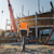 A construction worker in a hard hat and safety vest walks towards a building under construction with a large crane in the background.