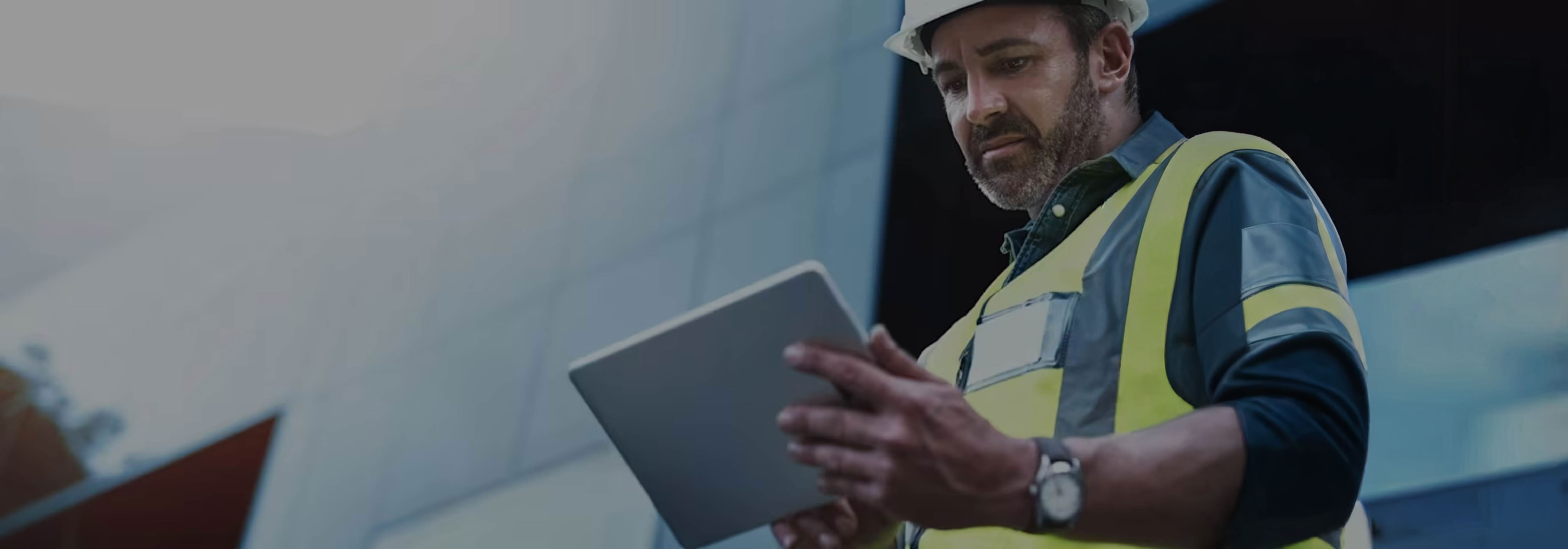A man wearing a white hard hat and a yellow high-visibility safety vest is shown outdoors, looking intently at a tablet he holds with both hands.