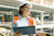 A woman in a hard hat and orange vest smiles while holding a tablet and papers at a construction site.