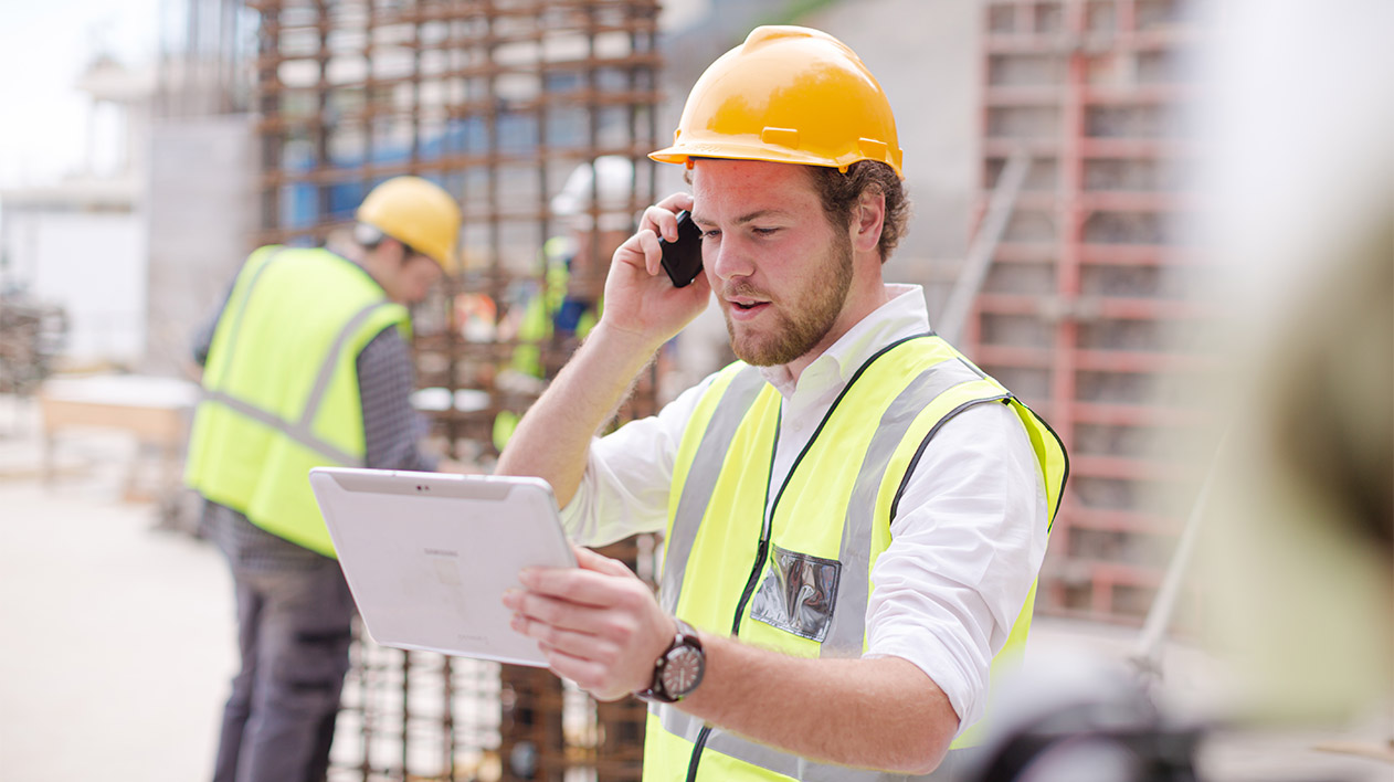 A man in a yellow hard hat and safety vest talks on a phone while holding a tablet at a construction site.