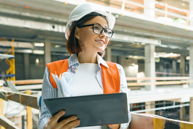 A woman in a hard hat and orange vest smiles while holding a tablet and papers at a construction site.