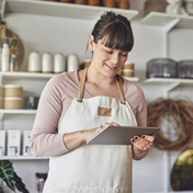 A smiling woman wearing an apron stands in a store, looking down at a tablet she is holding.