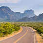 A winding two-lane road leads through a desert landscape toward the rugged Chisos Mountains and Casa Grande peak in Big Bend National Park, Texas.
