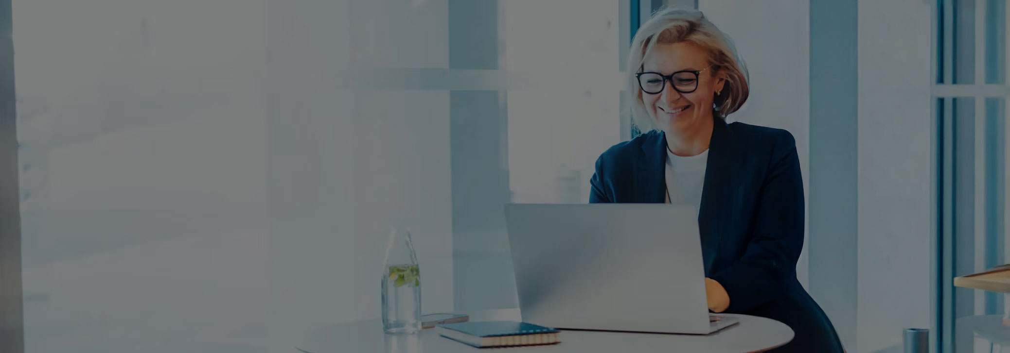 A smiling woman with short blonde hair and glasses sits at a table, looking at a laptop.