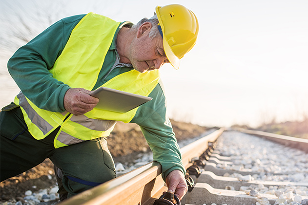 A worker in a yellow hard hat and vest inspects a train track while holding a tablet.