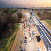 An aerial view shows highway construction next to active traffic, with cranes and heavy equipment.