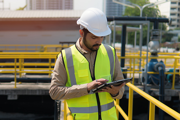  A man in a hard hat and safety vest uses a tablet at a water treatment facility with yellow railings.