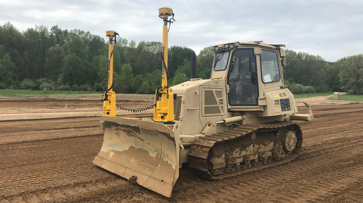 A bulldozer with Trimble Geospatial equipment, including two tall poles with sensors, sits on a dirt construction site with trees in the background.