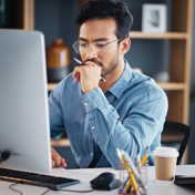 A professional-looking man with glasses and a blue collared shirt is sitting at a desk looking at a computer monitor.