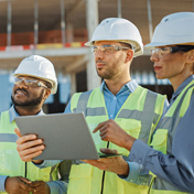 Three construction managers with hard hats and reflective vests looking at building and using laptop.