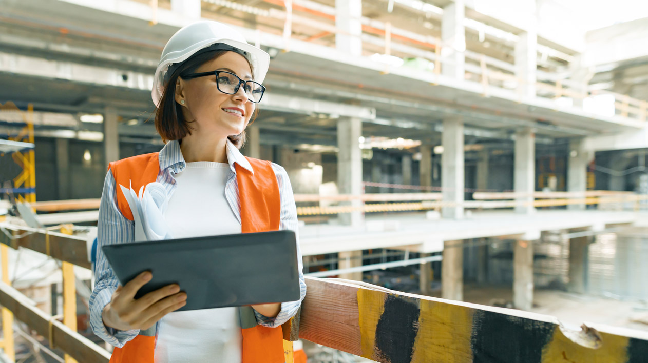 A woman in a hard hat and orange vest smiles while holding a tablet and papers at a construction site.