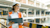 A woman in a hard hat and orange vest smiles while holding a tablet and papers at a construction site.