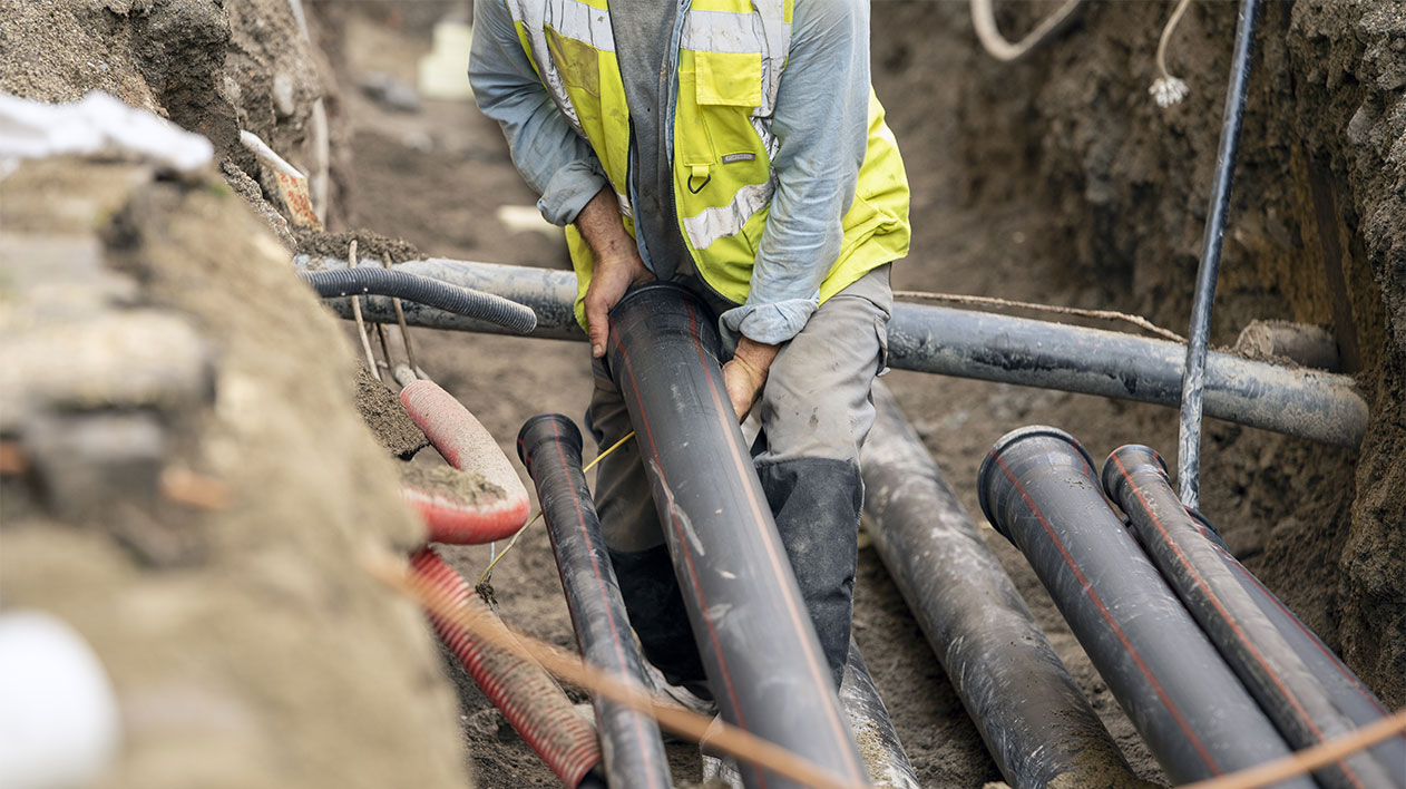 A construction worker in a safety vest and hard hat is in a trench, laying large black pipes and other conduits in the ground.