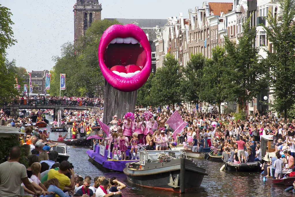 A group of people dressed in pink sailing through Amsterdam's canal. At the quay, there is a crowd of people cheering.