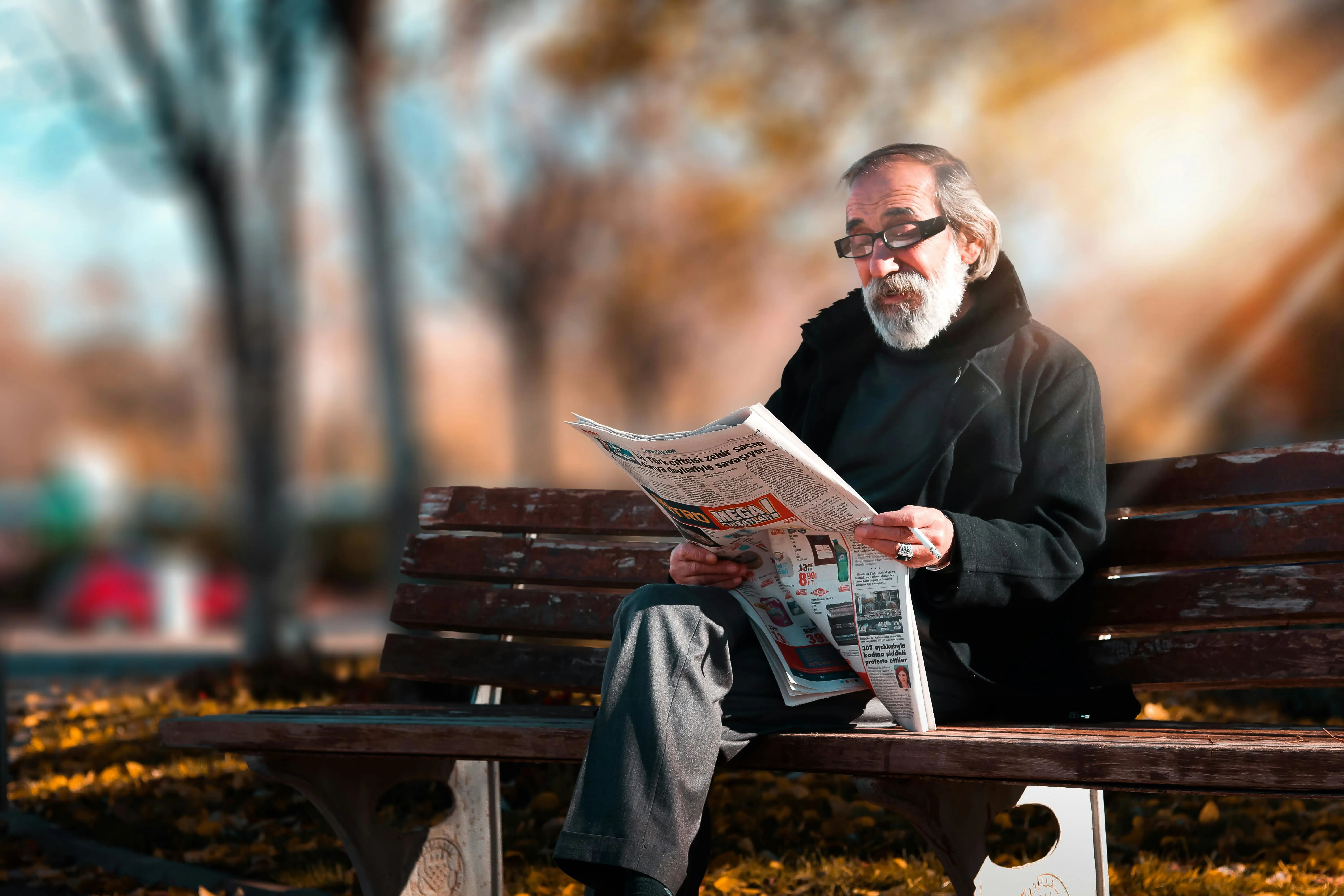 An elderly man sits on a bench reading the newspaper.
