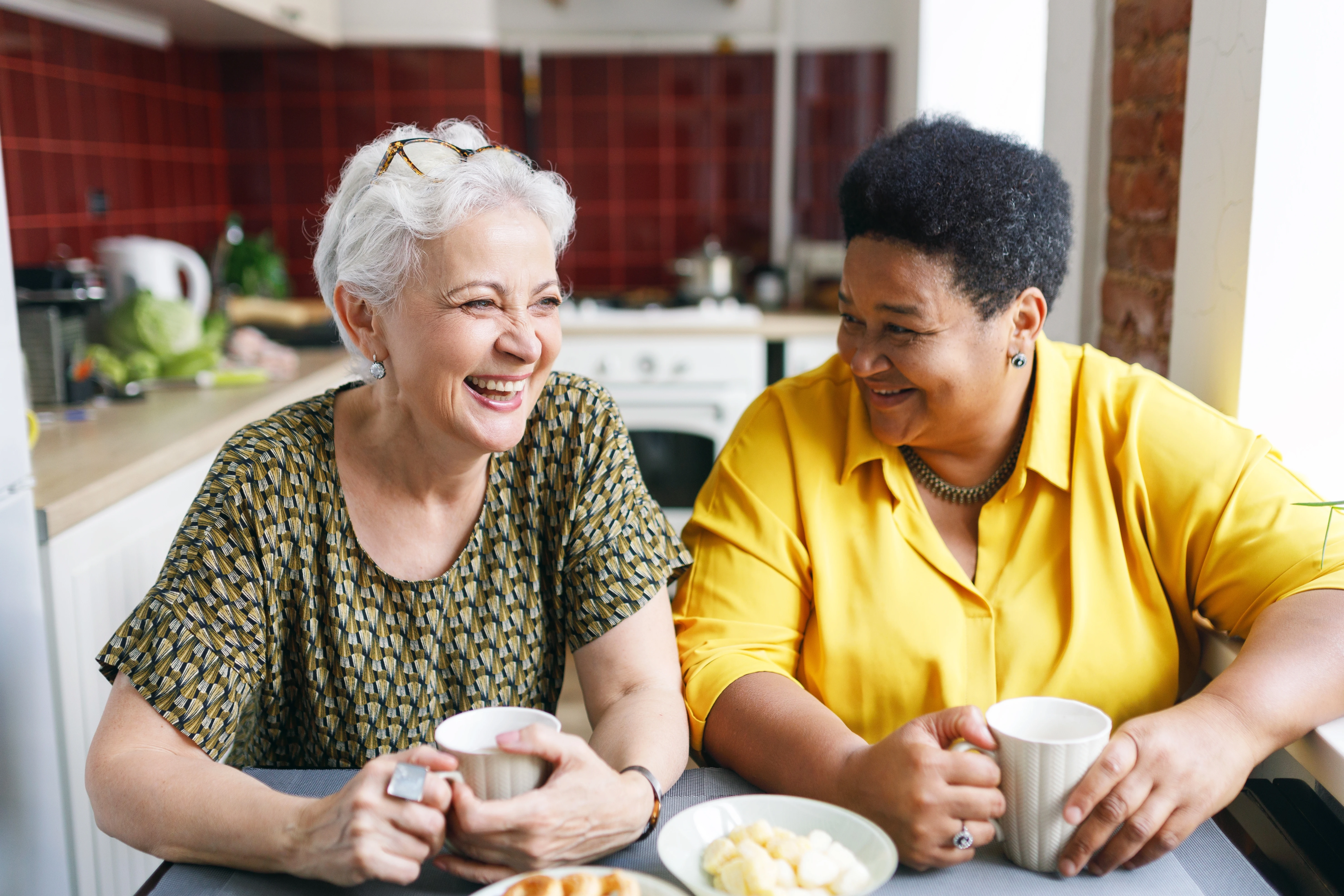 Twee aan een keukentafel zittende vrouwen lachen samen om een grappige situatie terwijl ze koffie met zoetigheid drinken