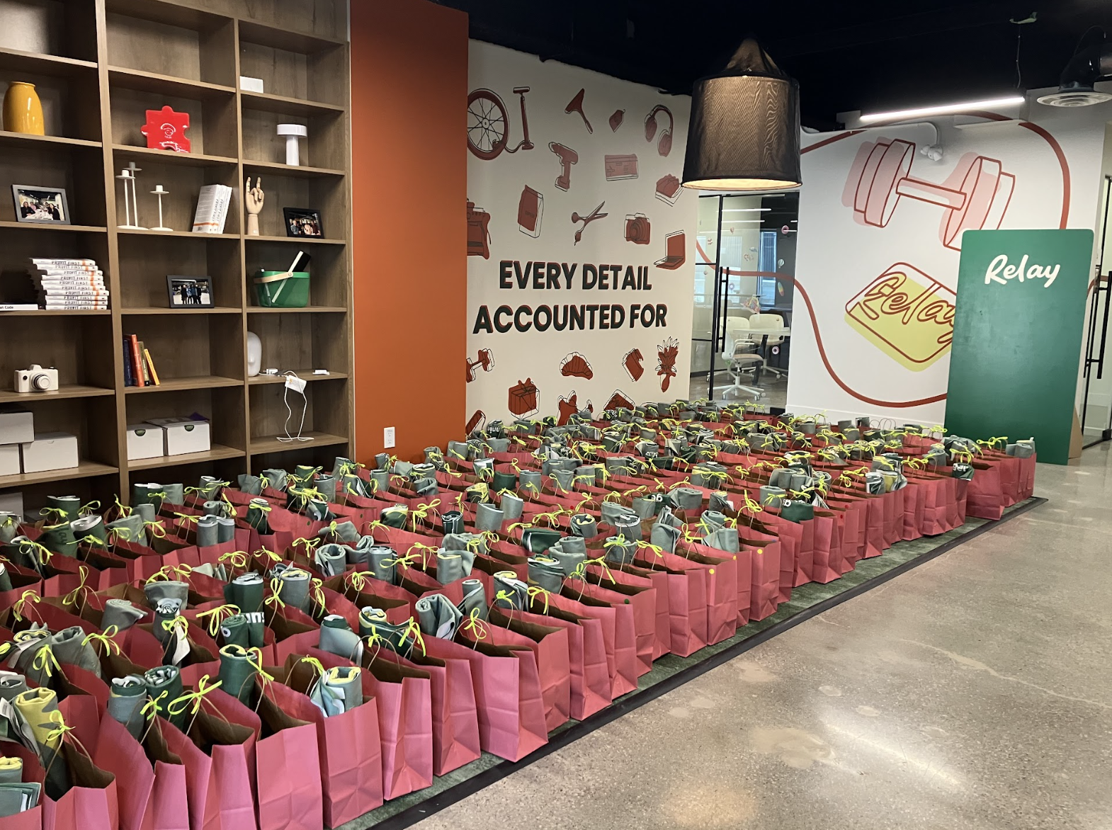 Lines of pink gift bags with green bows in an office. There are roughly 70 bags.