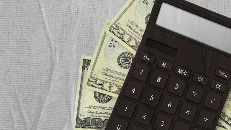 Black calculator resting on twenty dollar bills on a white textured surface with faint financial document visible.