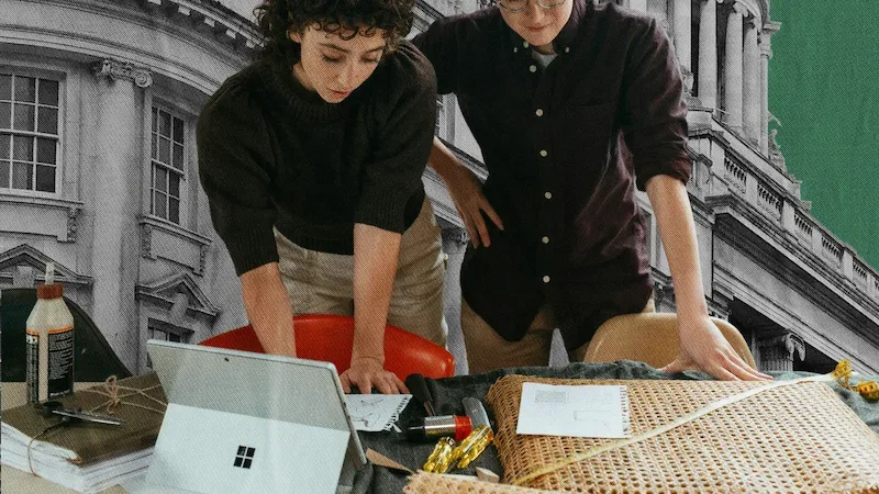 Two people in black shirts working together at a table with laptops and materials in front of a building backdrop.